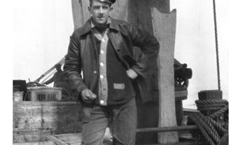 Black-and-white photo of a young man in a peaked cap, jacket and tall rubber boots. He stands on deck beside the mast with one hand on his hip, with coils of rope at his feet.