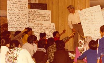 A man in a white shirt alongside some white signs with letters teaches a room full of children