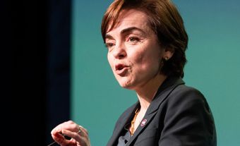 A woman with short hair wearing a dark blazer speaking at a microphone.