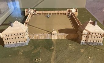Wooden scale model on fake grass behind a glass pane.