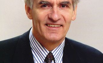 A smiling white man. He is wearing a dark suit, a tie, and a white shirt with blue stripes.