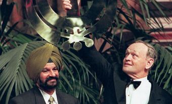 A man wearing a dark yellow turban clapping while a white man on the right holds up a Sikh symbol