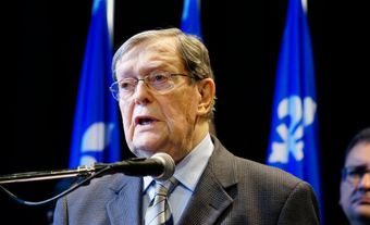 An elderly man with glasses in a grey suit. Three Quebec flags can be seen behind.