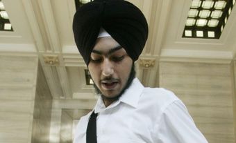 Un jeune homme portant un kirpan, un turban noir, une chemise blanche et un pantalon noir. Il se tient debout dans une grande pièce blanche.