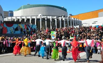 Des danseurs en habits colorés se tenant les uns aux autres en ligne. Ils présentent devant une foule dans un lieu public.