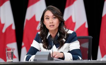 A woman with dark brown hair wearing a striped blue-white outfit is seated at a table with a microphone. Behind her is a row of Canadian flags.