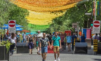People walking in the street with trees and signs on the side. There are multicoloured balls suspended above.