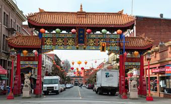 Une porte du quartier chinois avec un lion en pierre de chaque côté. Derrière, on aperçoit une rue où sont garés des voitures et des camions.